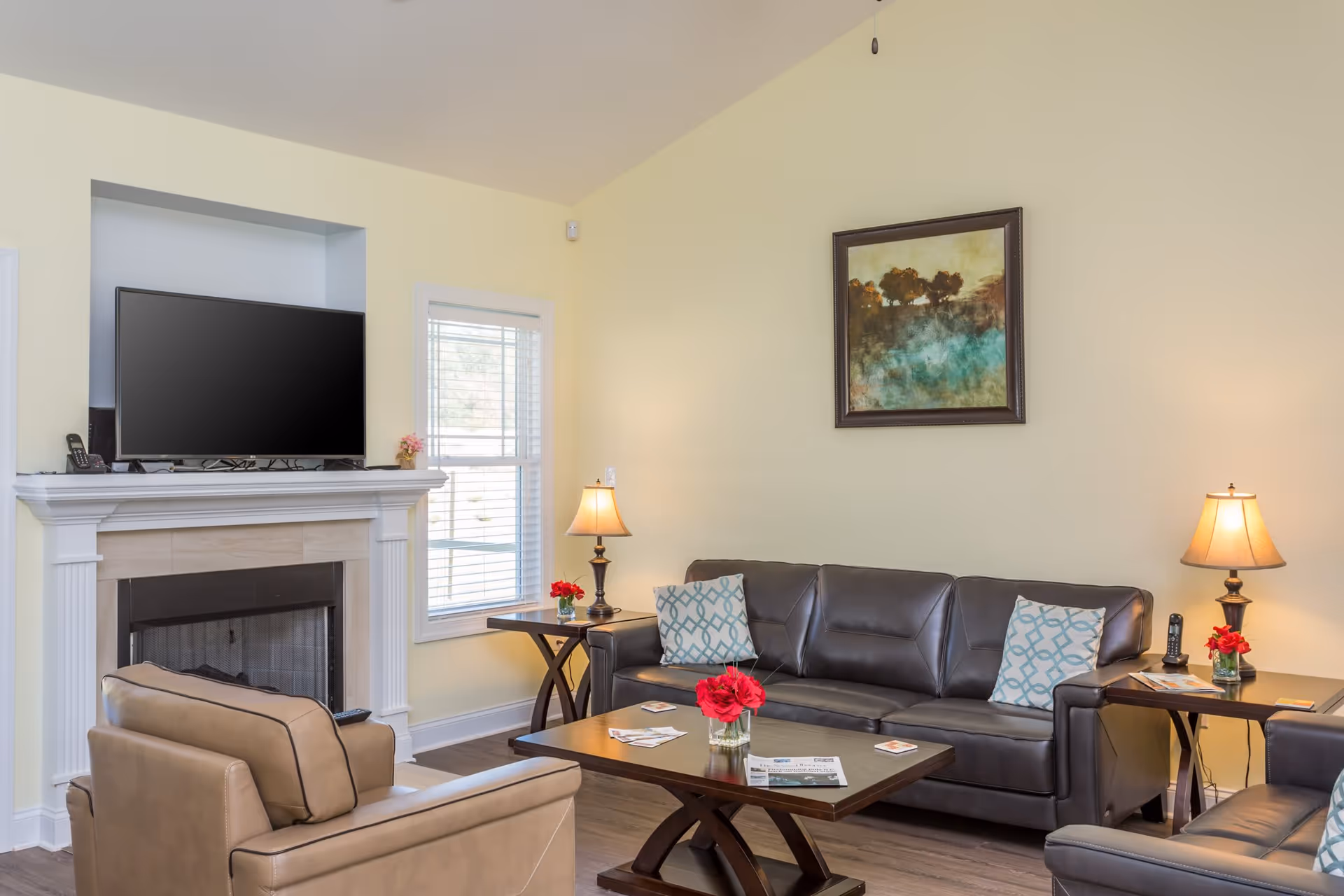 Living room with a leather sofa and armchairs arranged around a coffee table, a fireplace with a TV above, side tables with lamps, and a framed painting on the wall.