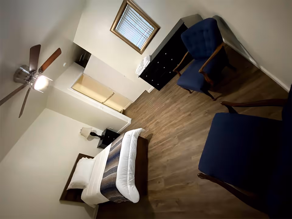 A bedroom with a bed made with white linens and a patterned blanket, two blue upholstered chairs with wooden arms, a black dresser with folded white towels on top, a nightstand with a lamp, a ceiling fan with a light, and a window with blinds and a gold frame mirror above the dresser.