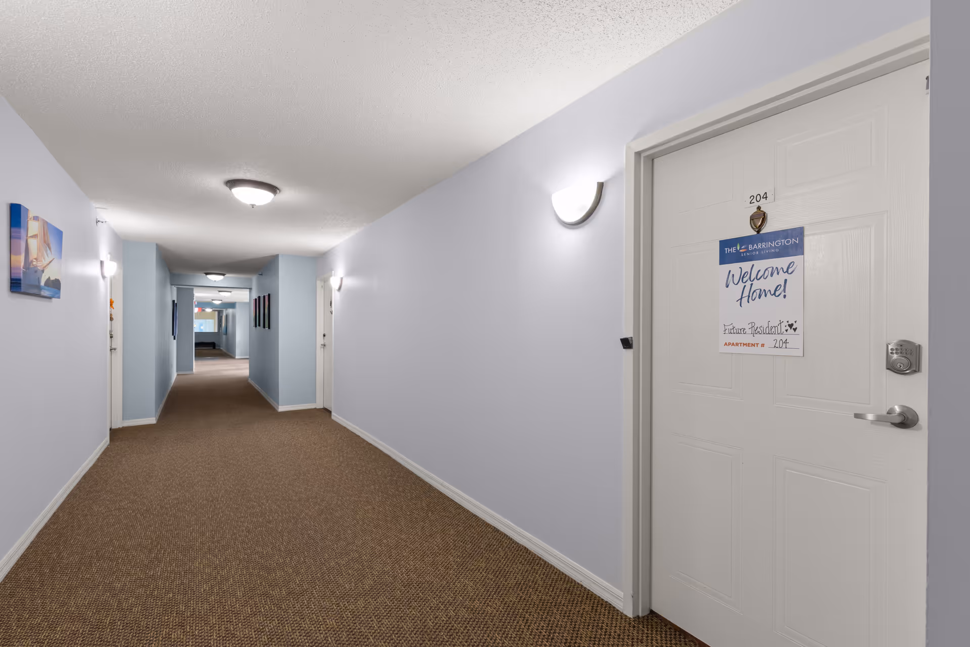 Interior hallway of The Barrington facility with light blue walls, brown carpet, and ceiling lights. A white door labeled 204 has a sign that reads 'Welcome Home! Future Resident Apartment 204'.