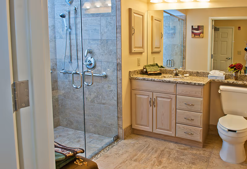 Light-colored bathroom with a glass-enclosed tiled shower, granite-top vanity with sink and cabinets, and a toilet.