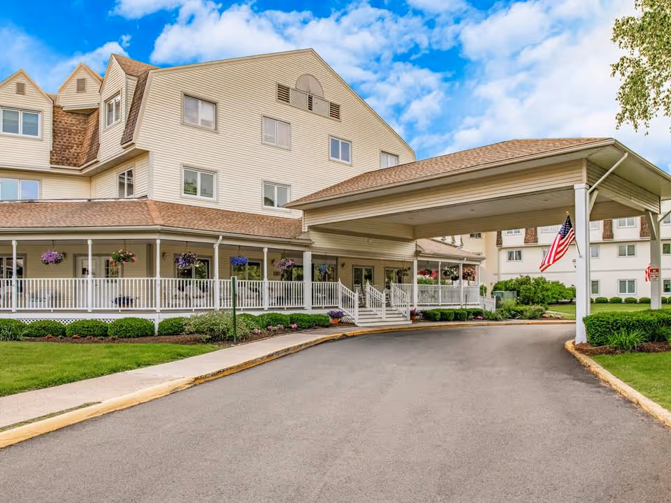Exterior view of Holiday Farmington senior living facility showing a multi-story building with beige siding, a covered entrance with white railings, hanging flower baskets, and an American flag. The driveway curves toward the entrance under a blue sky with scattered clouds.