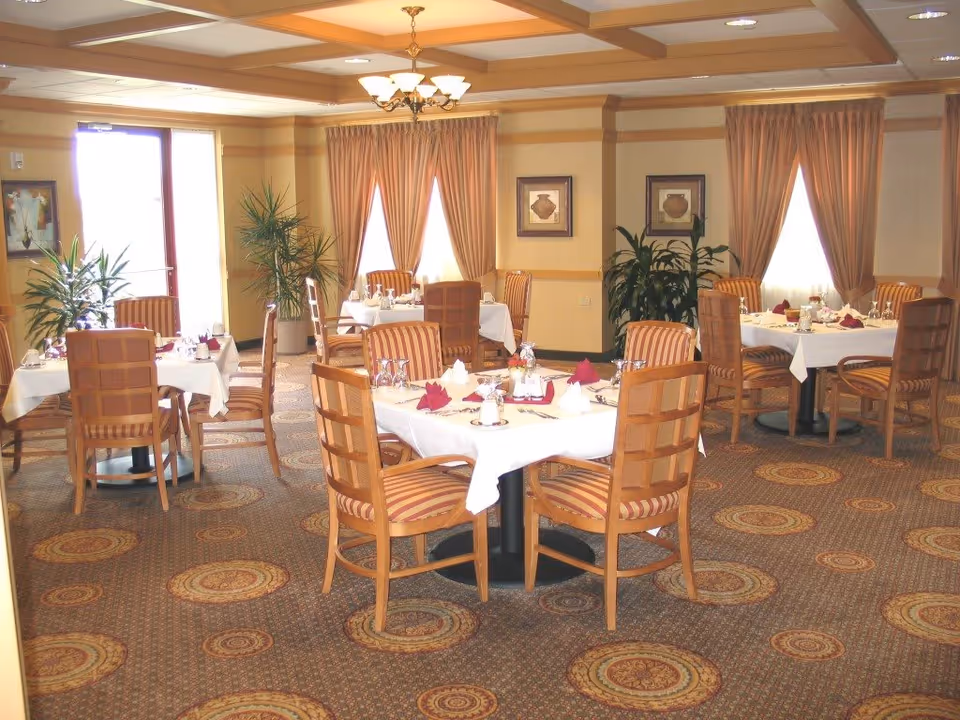 A dining room with several round tables covered with white tablecloths, each set with napkins, glasses, and silverware. The room has patterned carpet, striped upholstered chairs, large windows with peach-colored curtains, and potted plants near the walls. Warm lighting comes from a chandelier on the ceiling.