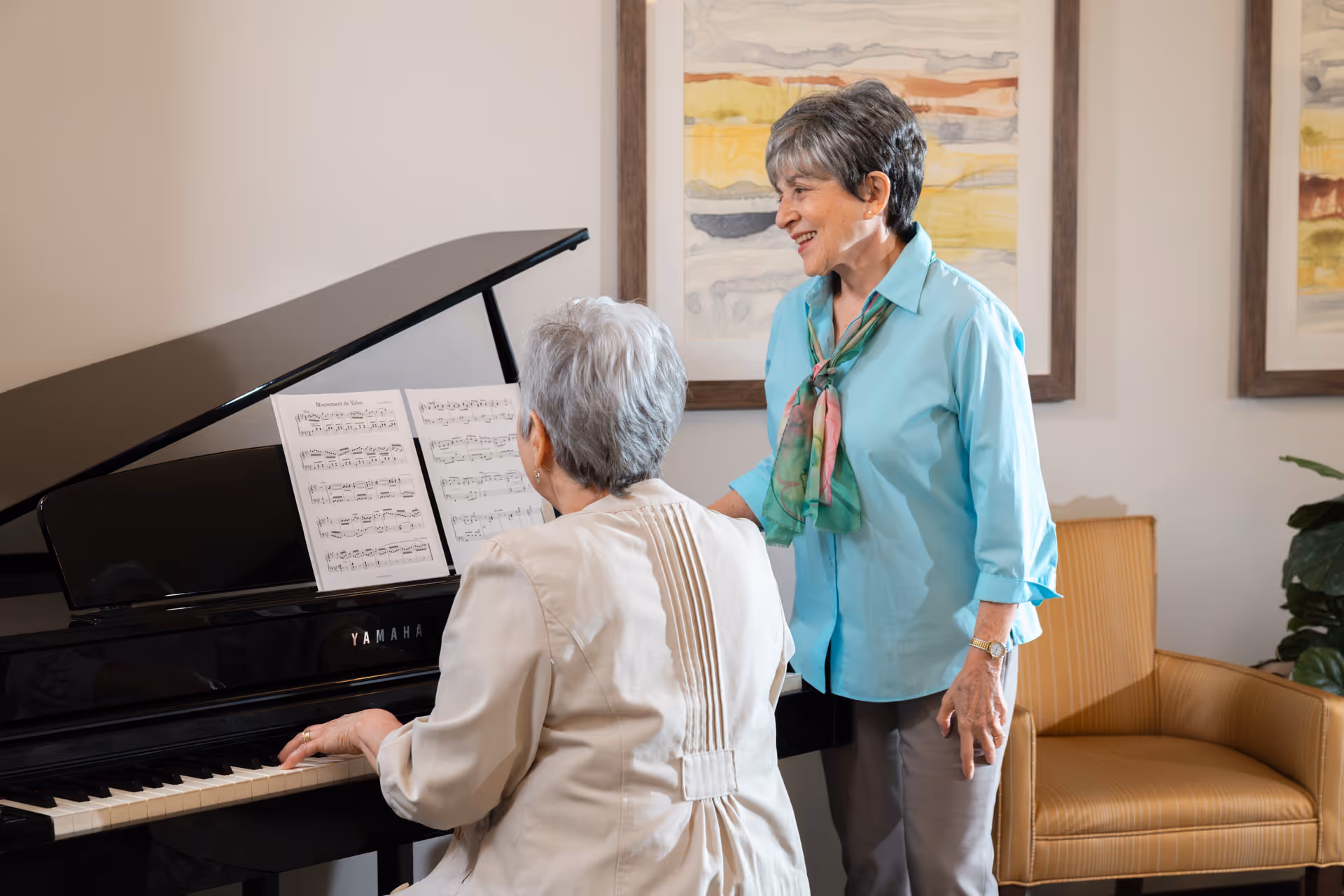 Two elderly women in a living room setting, one playing a black Yamaha piano with sheet music, and the other standing beside her smiling. The room has framed abstract artwork on the wall and a yellow armchair nearby.