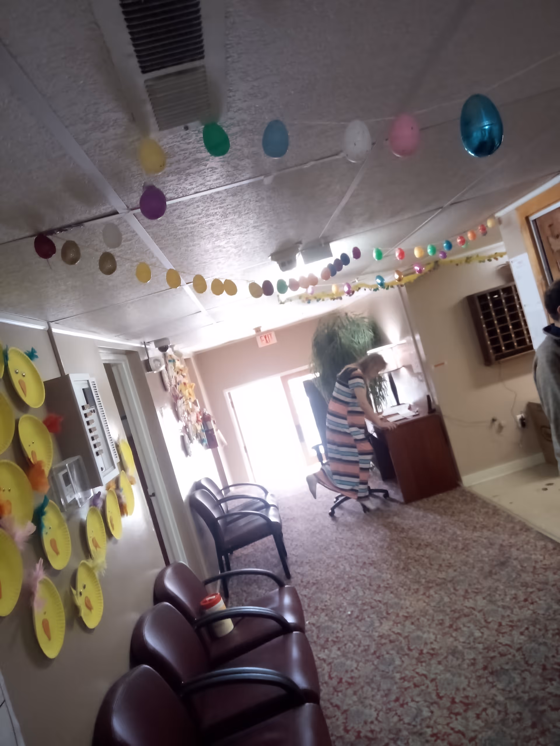 Interior hallway of a residential care facility decorated with colorful hanging Easter egg garlands and paper plate crafts resembling chicks on the wall. Several chairs line the left side of the hallway, and a woman in a striped dress is standing and leaning over a desk near the far end of the hallway. A large plant is visible behind the desk.