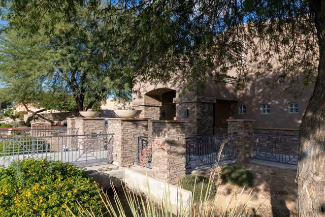 Stone-pillared entrance and landscaped front area of a building with metal railings and trees.
