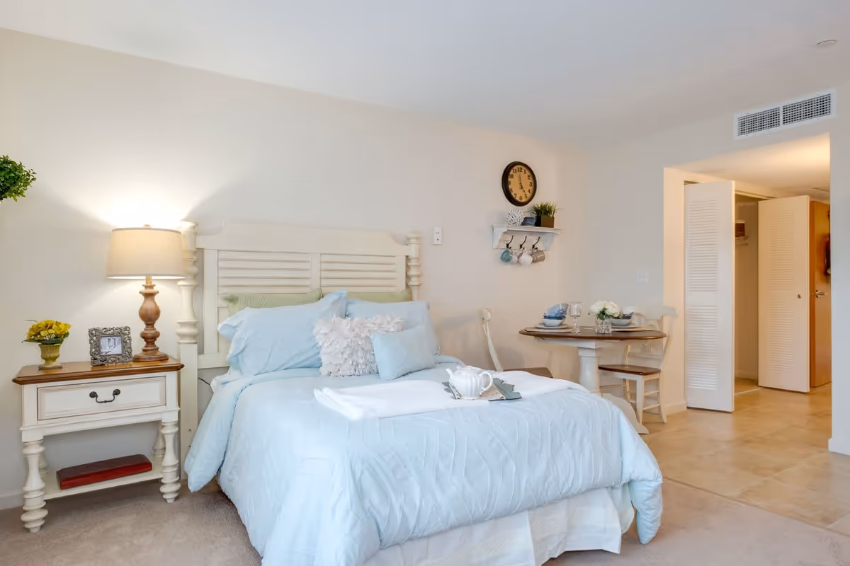 A cozy bedroom with a light blue bedspread and multiple pillows on a white wooden bed frame. To the left of the bed is a nightstand with a lamp, a small plant, and a framed photo. On the right side, there is a small round dining table set with plates, bowls, glasses, and a flower vase, accompanied by two chairs. A wall clock and a small shelf with hanging mugs are mounted on the wall above the dining table. The room has beige walls and carpeted flooring, with an open doorway leading to another area with tiled flooring.