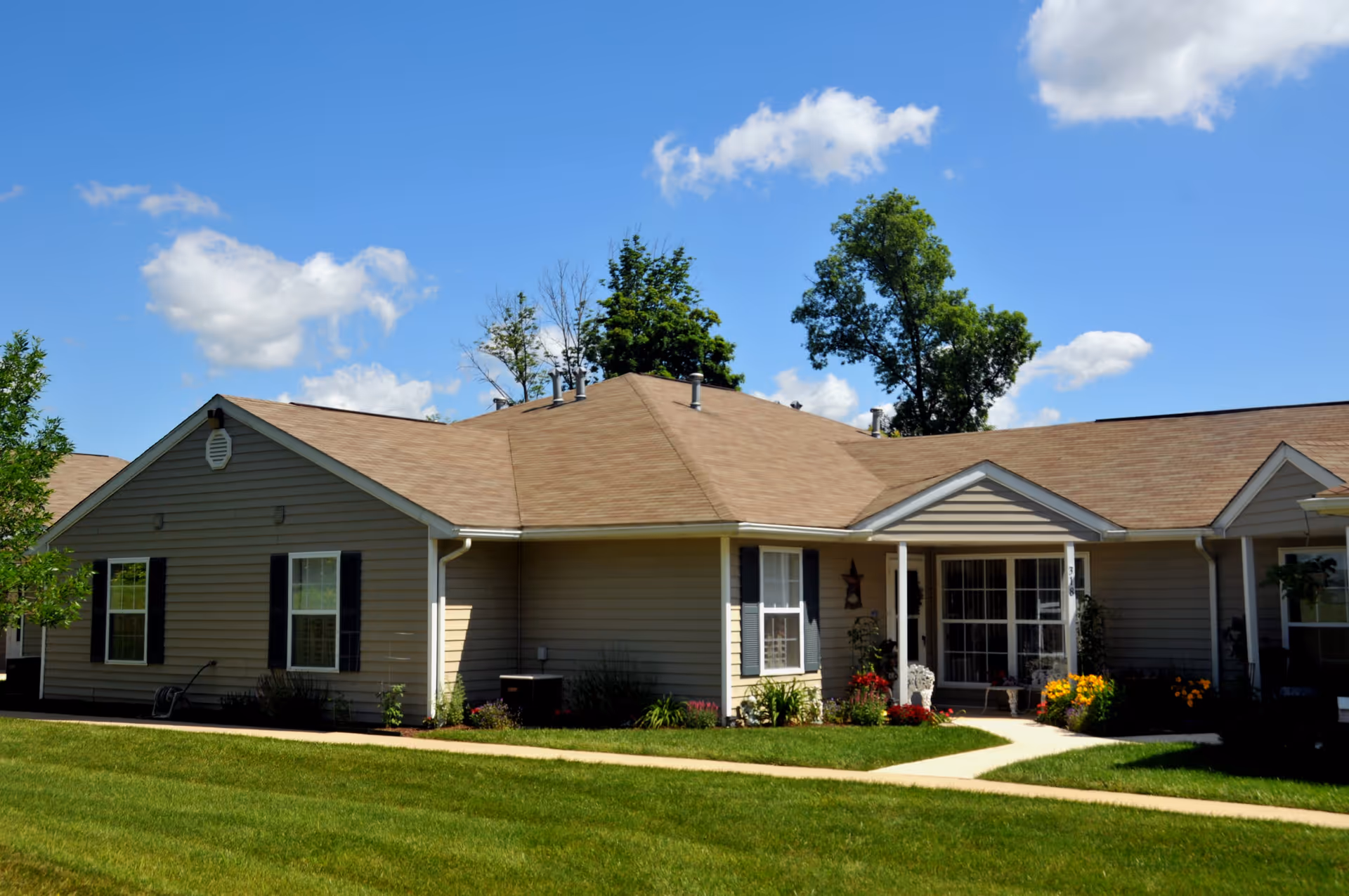 Single-story beige residential building with a brown shingled roof, white trim, and black window shutters. The building is surrounded by a well-maintained green lawn, a concrete walkway, and colorful flower beds near the entrance. Trees and a blue sky with scattered clouds are visible in the background.
