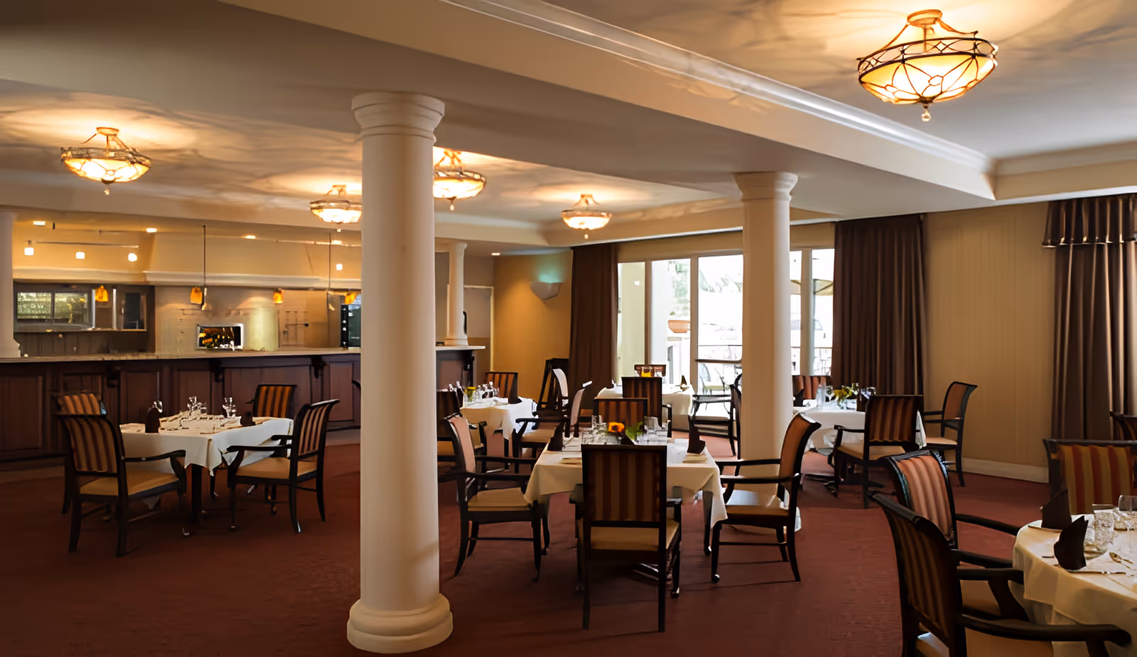 Spacious dining room with tables set for meals, upholstered chairs, decorative columns and warm ceiling lights.