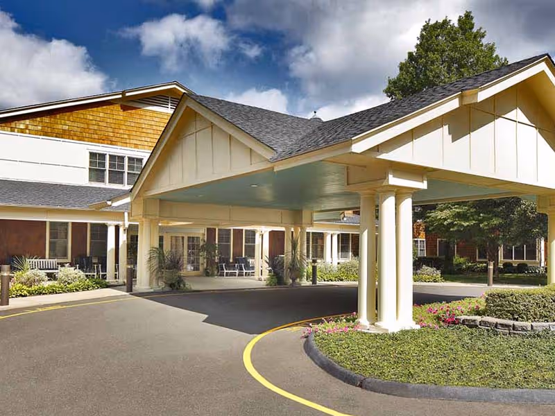 Entrance of a senior living facility with a covered drop-off area supported by white columns, surrounded by landscaped greenery and flowers under a partly cloudy sky.