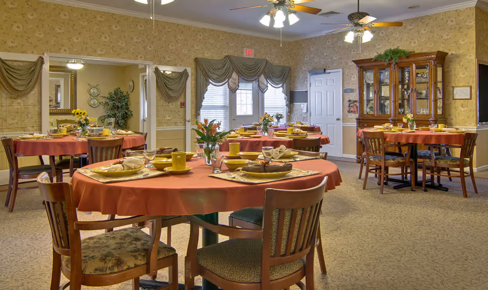 A dining room in a senior living facility with round tables covered in red tablecloths, each set with yellow cups, plates, napkins, and silverware. The room has floral wallpaper, carpeted floors, wooden chairs with patterned cushions, a wooden china cabinet, and windows with draped curtains. Ceiling fans with lights are visible above.