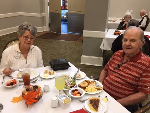 Two people sit at a dining table with plates of breakfast food, salads and drinks in a communal dining room.