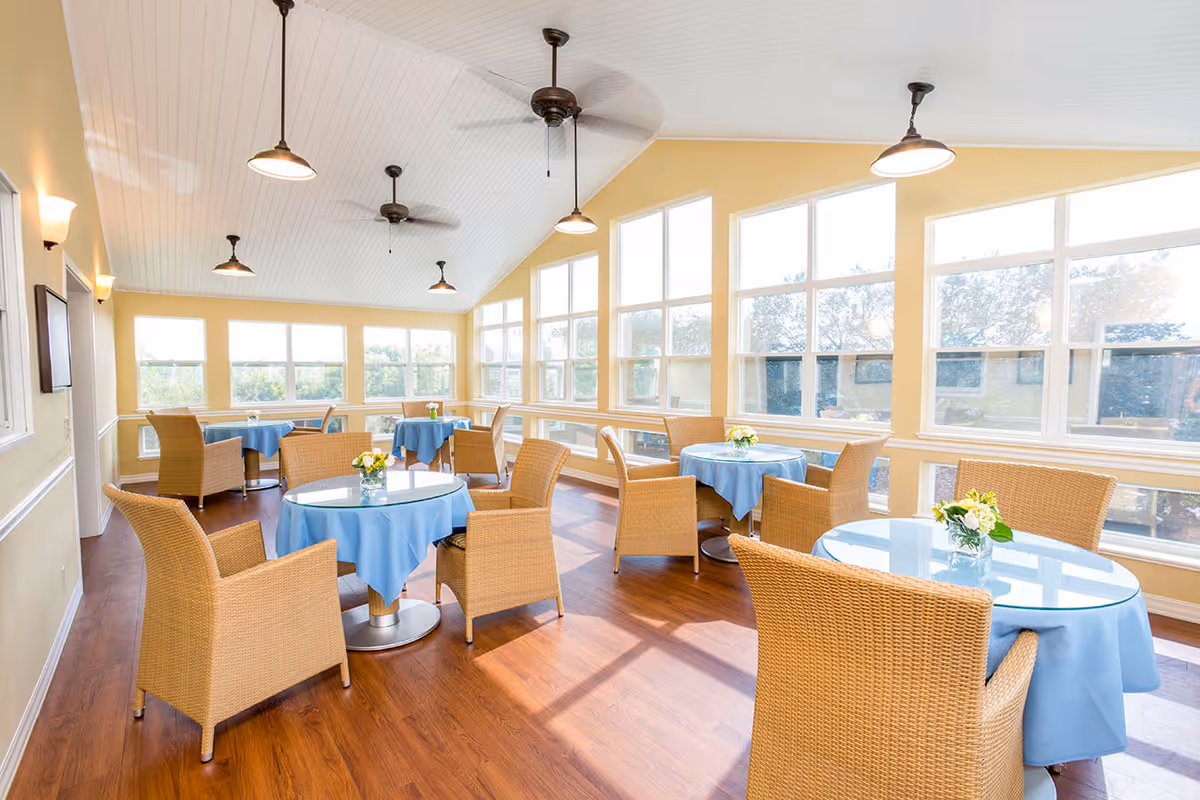 Bright dining room with multiple round tables covered with blue tablecloths, each surrounded by wicker chairs. The room has large windows letting in natural light, wooden flooring, ceiling fans, and pendant lights hanging from a white paneled ceiling.