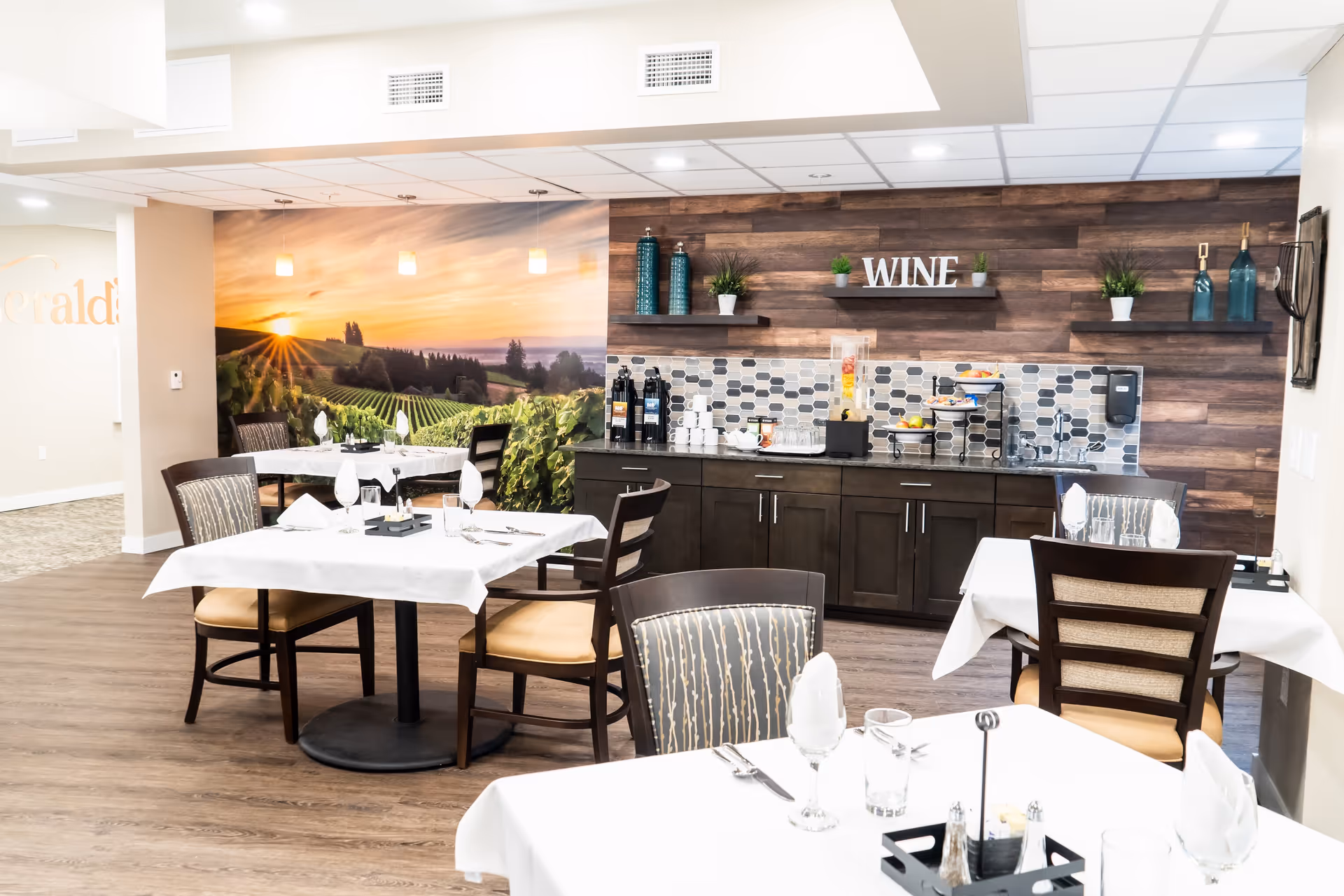 Bright dining area with white-clothed tables and chairs facing a wood-paneled beverage and wine service counter beneath a vineyard mural.