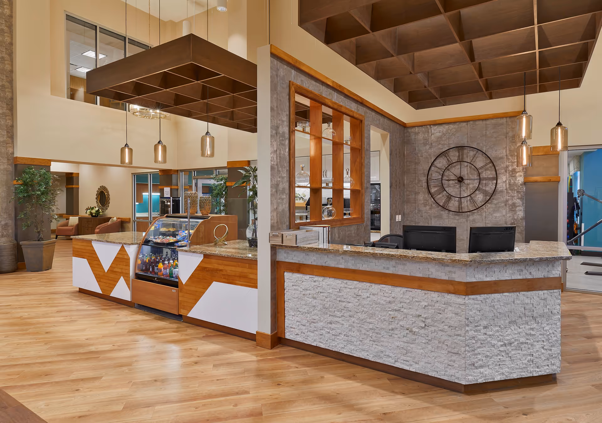 A modern reception area in a nursing home featuring a wooden and stone front desk with two computer monitors, a display case with beverages and snacks, pendant lights hanging from the ceiling, a large decorative wall clock, and seating area with plants and a mirror in the background.