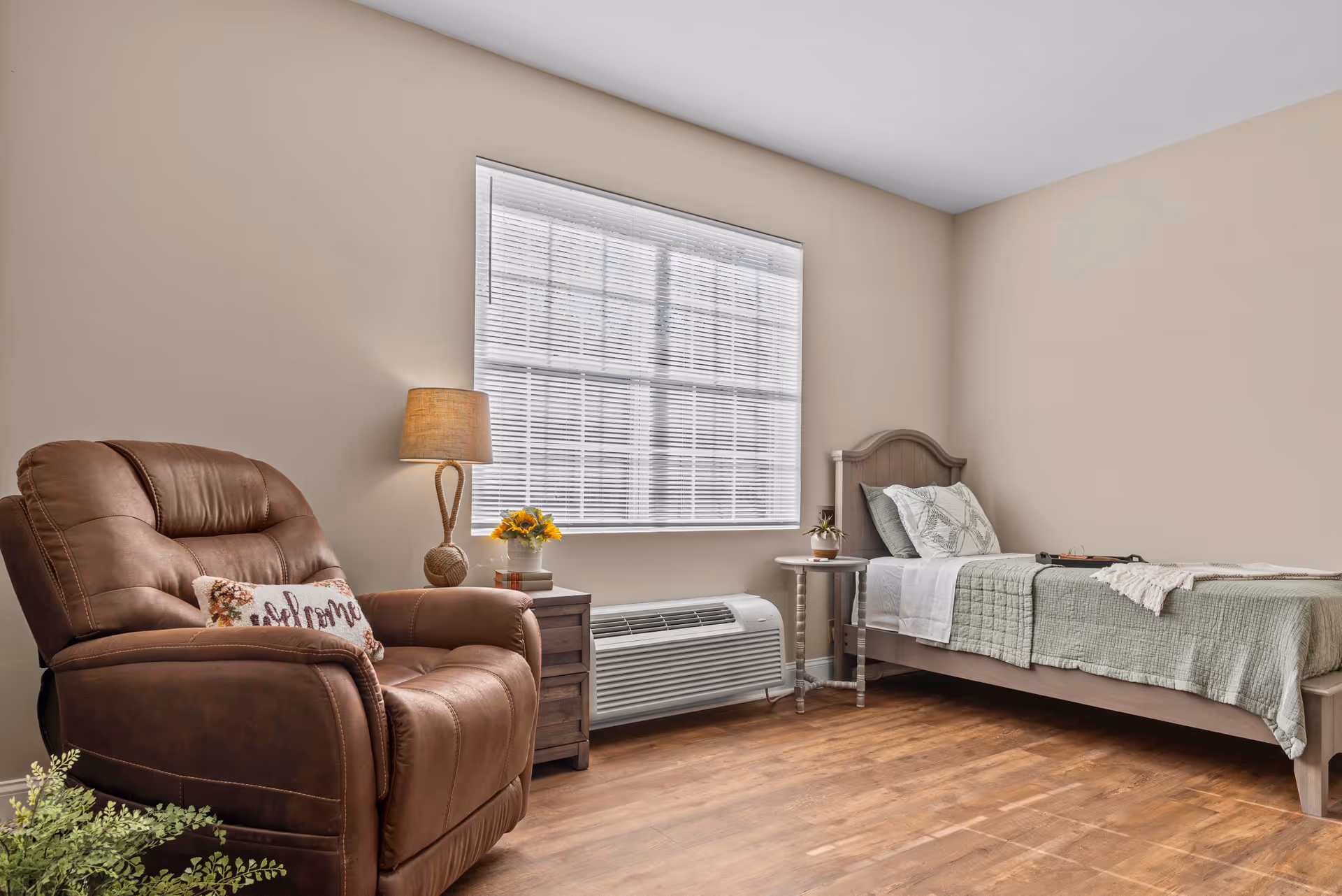 A cozy bedroom with a single bed covered in green and white bedding, a wooden headboard, and a small round side table with a plant. Next to the bed is a wooden nightstand with a lamp and a small flower arrangement. A large brown leather recliner chair with a decorative pillow is positioned near the window with white blinds. The room has light beige walls and wooden flooring.