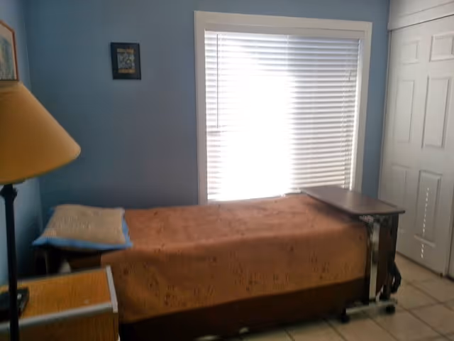 A small bedroom with a single bed covered with a brown blanket and a pillow. There is a window with closed white blinds behind the bed. A bedside table with a lamp is visible on the left side, and a small overbed table is positioned on the right side of the bed. The walls are painted blue, and there is a white door on the right side of the room.