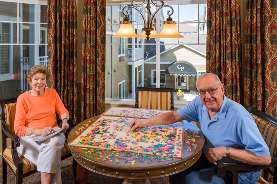 An elderly man and woman sitting at a round wooden table working on a large colorful jigsaw puzzle in a well-lit room with large windows and patterned curtains. The man is smiling and placing a puzzle piece, while the woman is holding a newspaper and smiling. Outside the window, a building with a canopy labeled 'Cp' is visible.