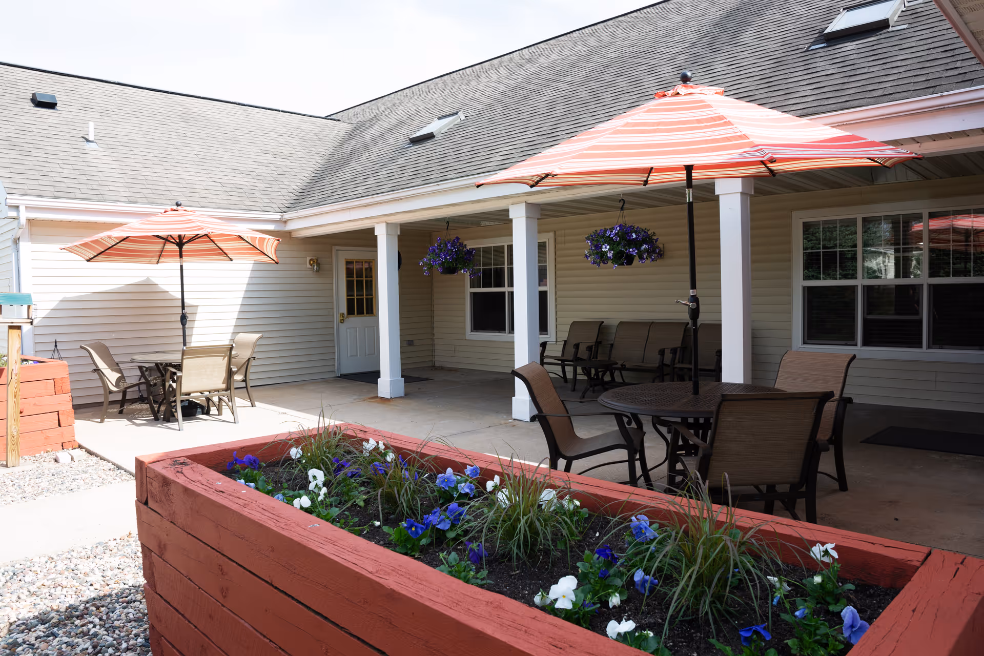 Shaded courtyard patio with tables, umbrellas, seating, hanging flower baskets and a red wooden planter in front of a beige building.