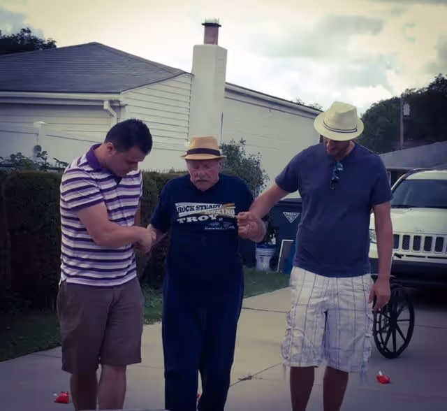 An elderly man wearing a hat is being supported by two younger men, one on each side, as they walk together outside on a driveway near a white building and a parked vehicle.
