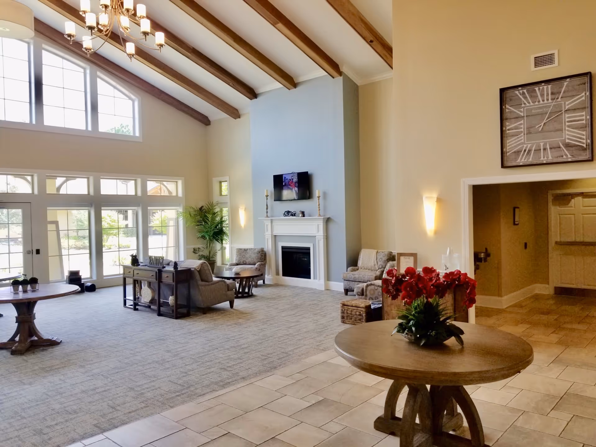 Spacious living room area with high vaulted ceiling featuring wooden beams, large windows letting in natural light, a fireplace with a mounted TV above it, several armchairs, round wooden tables with flower arrangements, and a large wall clock above a doorway.