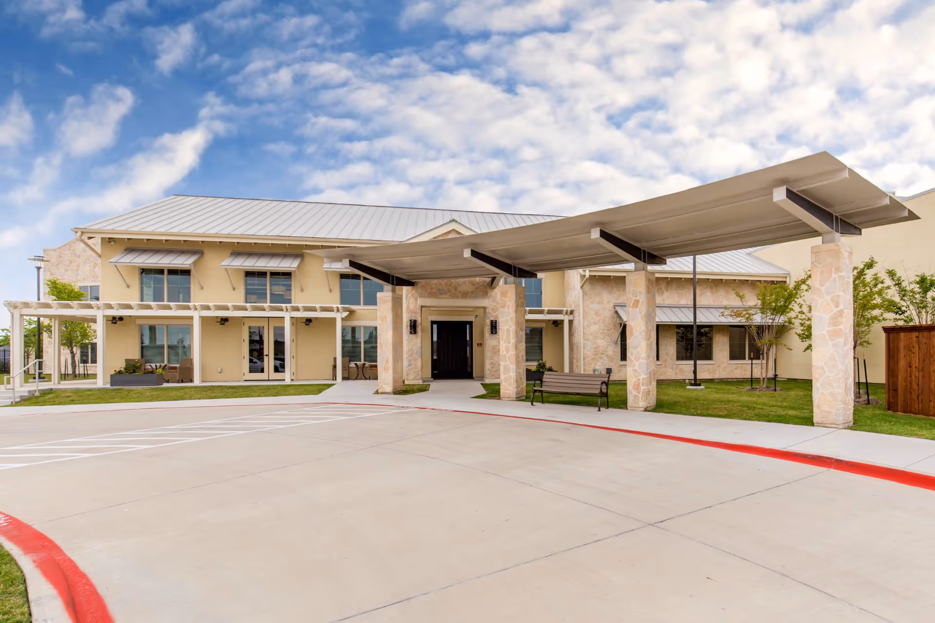 Front exterior view of a two-story senior living facility building with a covered entrance supported by stone pillars, a bench near the entrance, and a driveway with red curb markings. The building has a light yellow facade, multiple windows with awnings, and a metal roof under a partly cloudy sky.