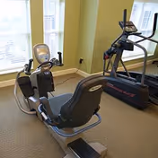 A small fitness room with exercise equipment including a recumbent bike and a treadmill, positioned near windows with natural light coming in.