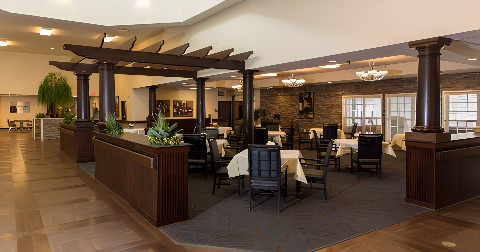 Interior view of a senior living facility dining area with multiple tables covered with white tablecloths and surrounded by chairs. The space features dark wooden columns and a decorative wooden pergola structure. There are plants on the divider and a stone accent wall with artwork in the background. The floor is a combination of wood and carpet.