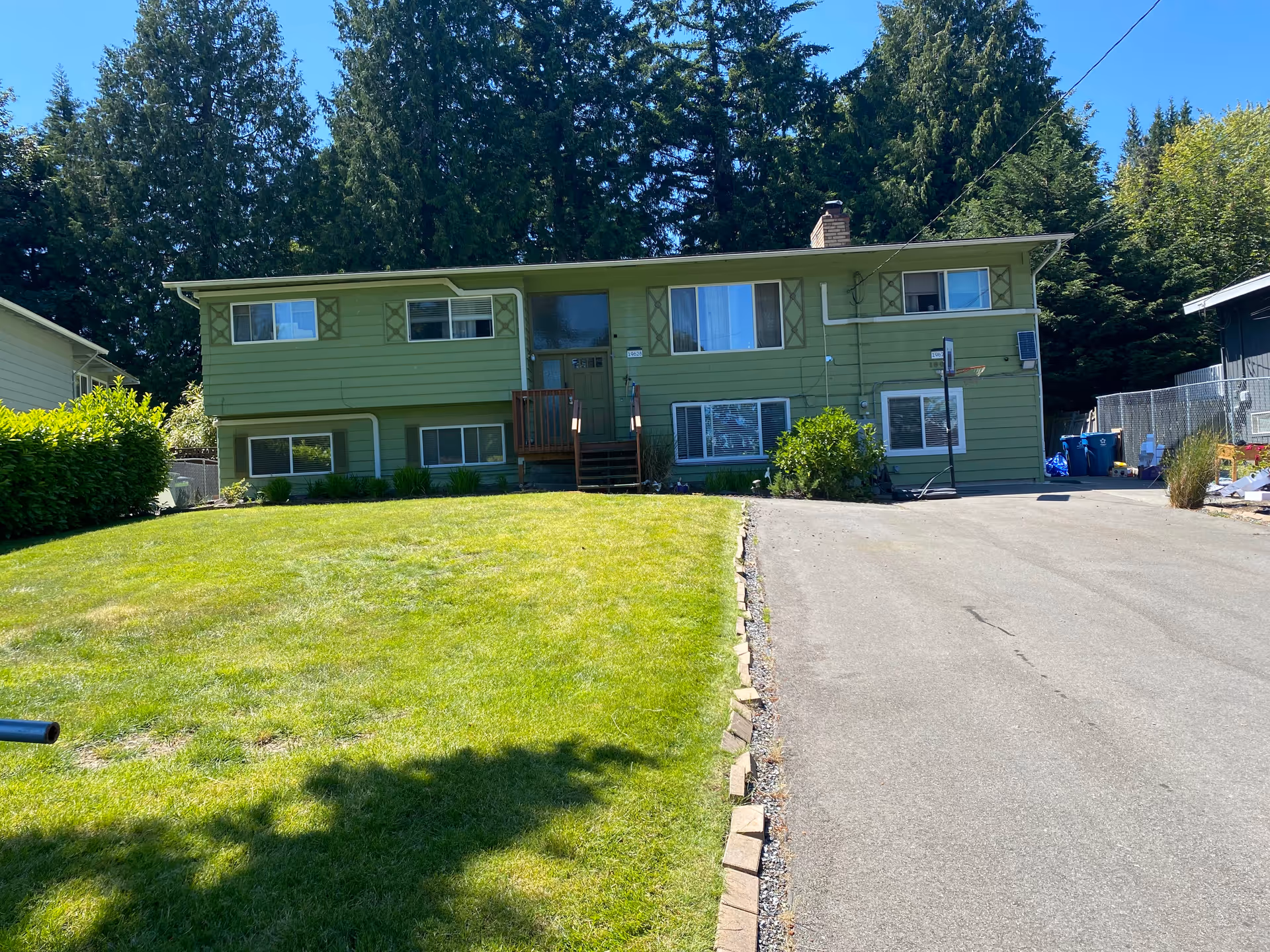 A green two-story house with multiple windows, a small front porch with stairs, a driveway on the right side, and a well-maintained lawn in front. Tall trees are visible in the background under a clear blue sky.