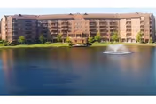 A large multi-story residential building with balconies overlooking a pond with a central water fountain, under a clear sky.