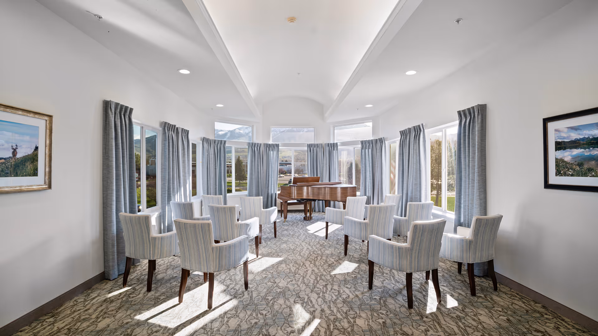 A bright room with large windows covered partially by gray curtains, arranged with a semi-circle of striped upholstered chairs facing a wooden grand piano. The room has a high vaulted ceiling with recessed lighting and framed landscape photographs on the walls.