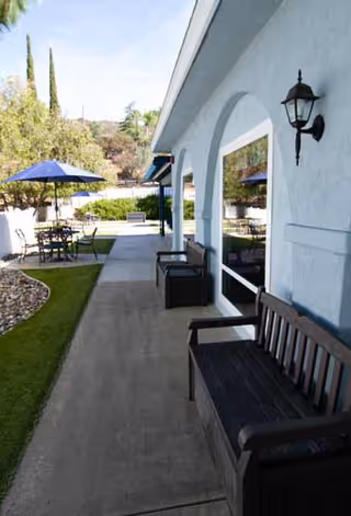 Outdoor patio area at Alpine View Lodge featuring benches along a building wall, a paved walkway, tables with umbrellas, and greenery including trees and bushes.