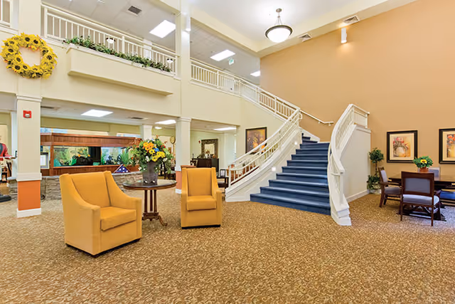 A spacious senior living facility common area with beige patterned carpet, two mustard yellow armchairs around a small round table with a flower arrangement, a blue-carpeted staircase with white railings leading to an upper floor, and a seating area with a table and chairs against a tan wall decorated with framed artwork and plants.