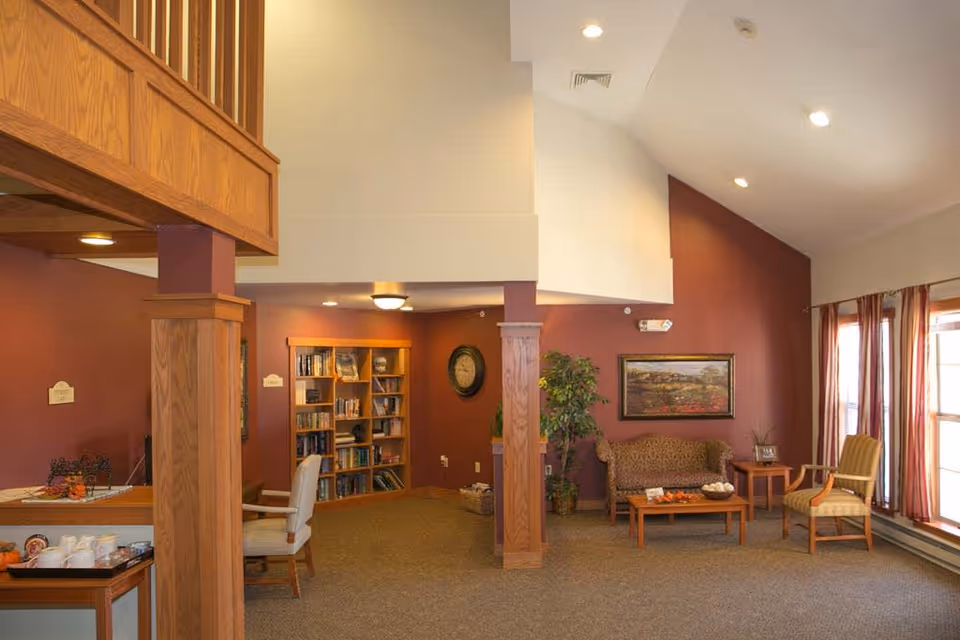 A cozy senior living facility common area with warm lighting and earth-toned walls. The space features a wooden bookshelf filled with books, a clock on the wall, a patterned sofa, two armchairs, a coffee table with decorative items, a side table with a framed photo, and large windows with striped curtains allowing natural light to enter. Wooden pillars and trim add to the warm ambiance.