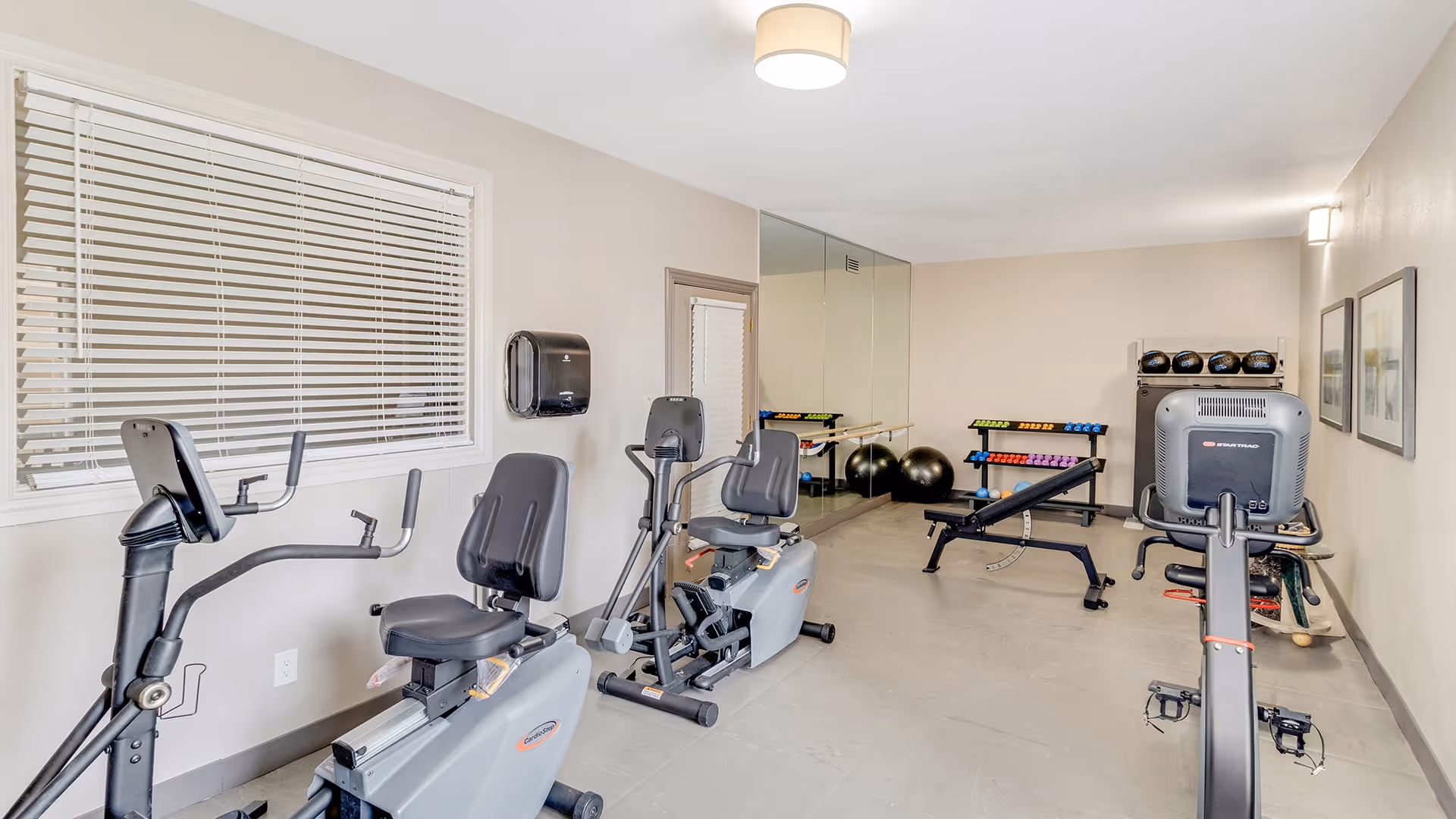 A small fitness room with two recumbent exercise bikes, a weight bench, a rack of colorful dumbbells, exercise balls, and medicine balls. The room has light-colored walls, a large window with blinds, a wall-mounted paper towel dispenser, and a large mirror on one wall.
