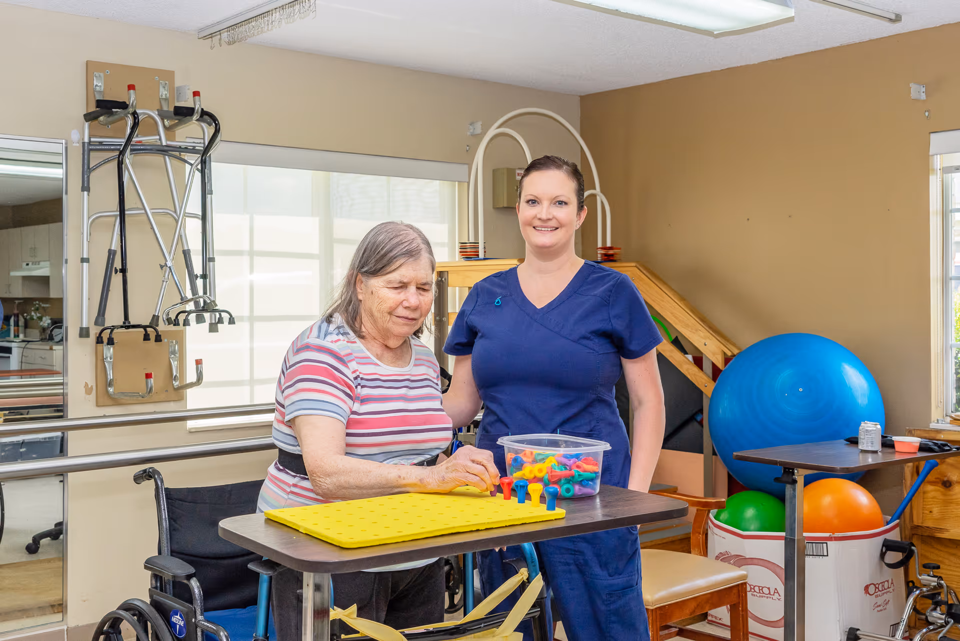 An elderly woman in a wheelchair is engaged in a therapeutic activity with colorful pegs on a yellow pegboard, assisted by a smiling female caregiver in blue scrubs. The room contains physical therapy equipment including exercise balls, parallel bars, and a walker mounted on the wall, with natural light coming through the windows.