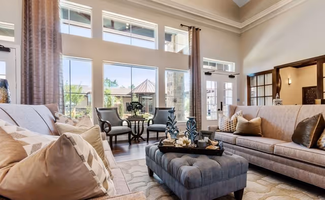 Sunlit communal living room with sofas, armchairs, a tufted ottoman and large windows overlooking a courtyard.