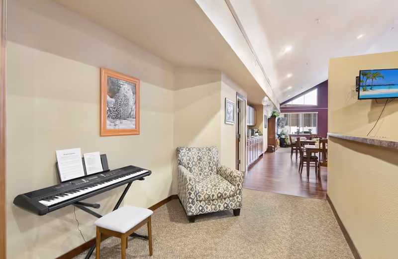 A bright interior common area with an electronic keyboard and patterned armchair in the foreground and a dining area with tables and chairs in the background.