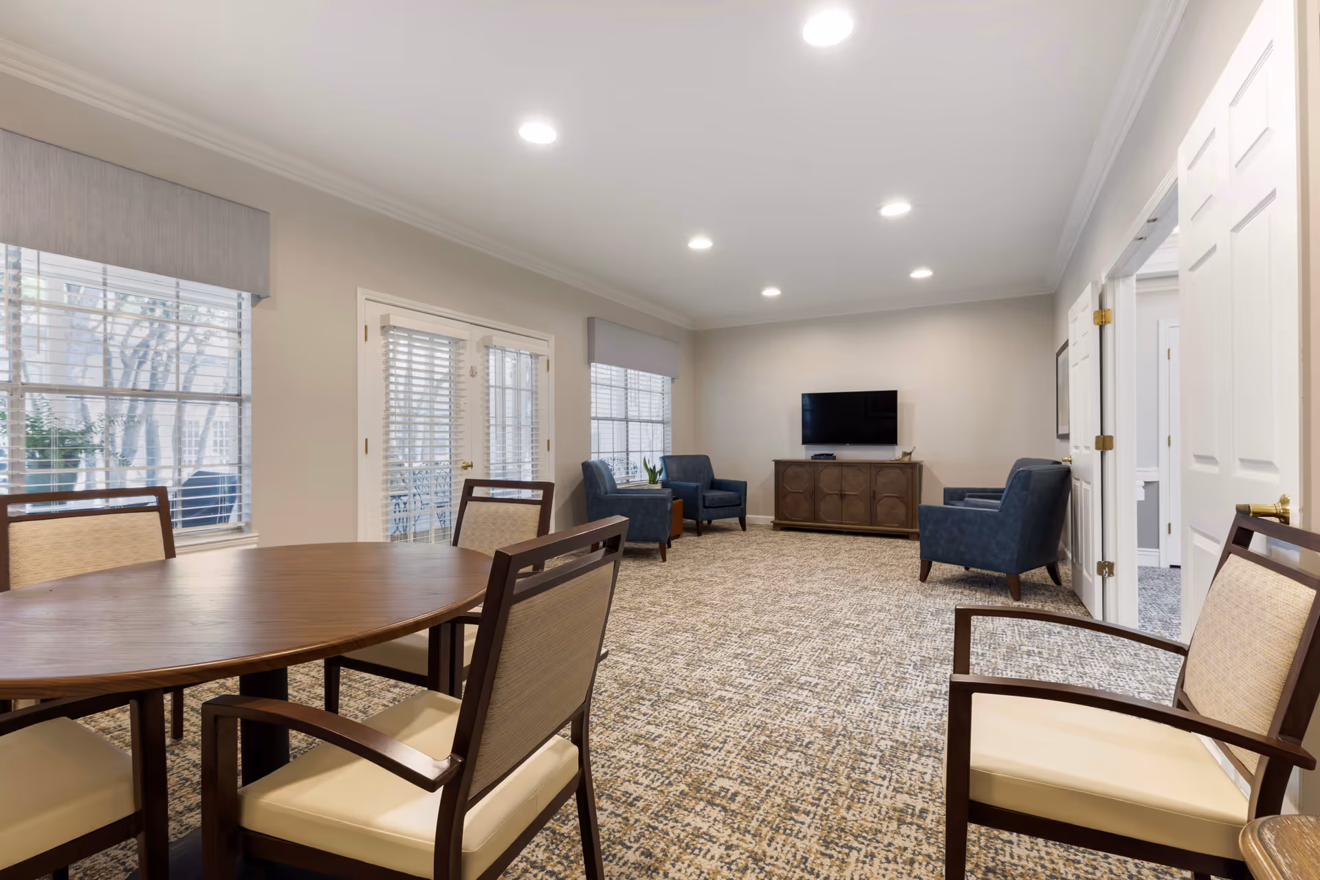 A spacious senior living common area with a round wooden table and four cushioned chairs in the foreground. The room features patterned carpet, beige walls, and multiple windows with blinds and valances. At the far end, there are three blue armchairs arranged around a wooden cabinet with a flat-screen TV mounted above it. Recessed ceiling lights illuminate the room.