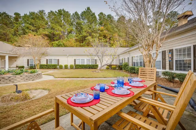 Outdoor patio area at The Legacy of Orangeburg with a wooden table set for six, featuring blue glasses and patterned plates on red placemats. The patio is surrounded by a grassy courtyard with trees and shrubs, and single-story building wings with white siding and multiple windows.