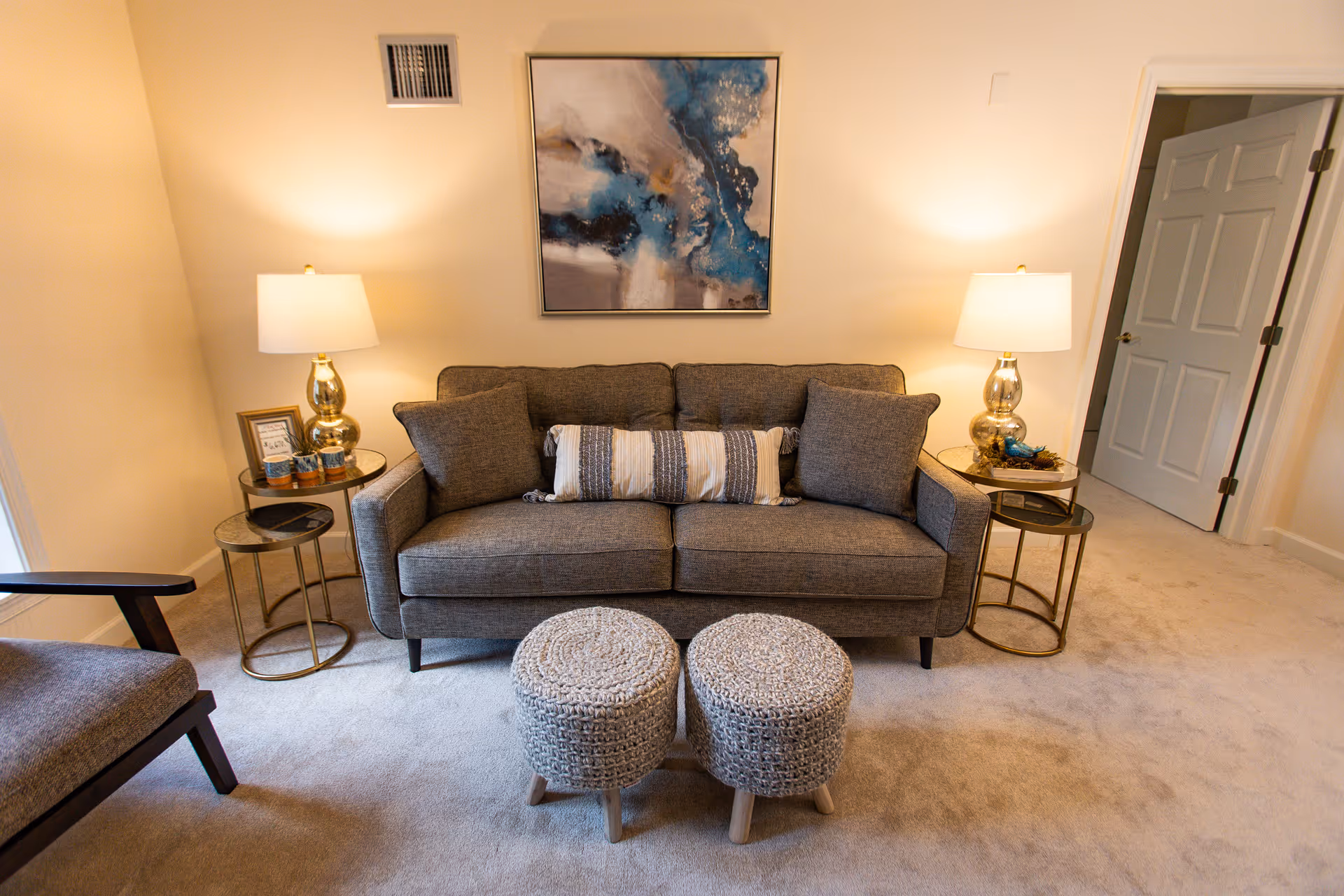 A cozy living room with a gray sofa, two woven ottomans, side tables with lamps, and abstract wall art.