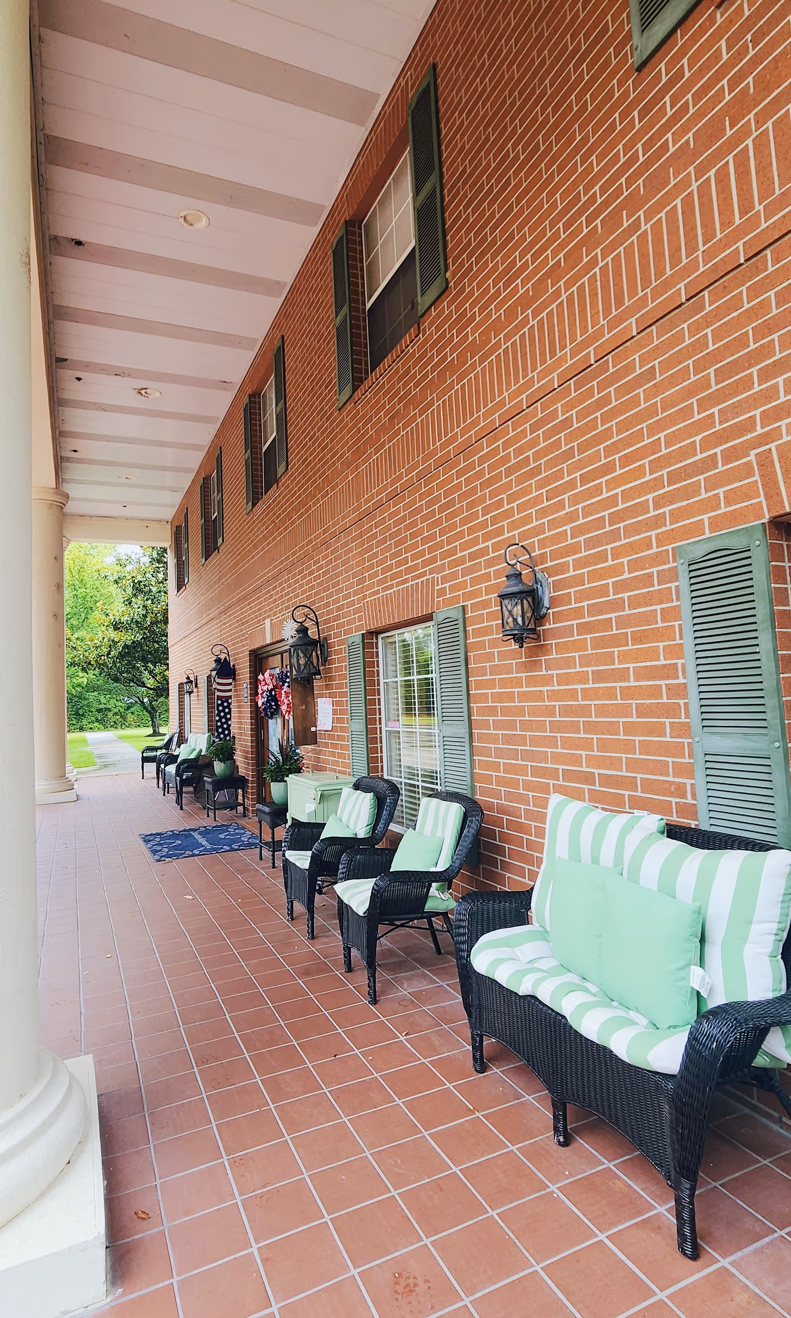 A covered outdoor porch area with a brick wall and several black wicker chairs and a loveseat with green and white striped cushions. The porch has a tiled floor and white columns, with green shutters on the windows and decorative wall lanterns. There are plants and a wreath on the door, and a grassy area with trees is visible in the background.