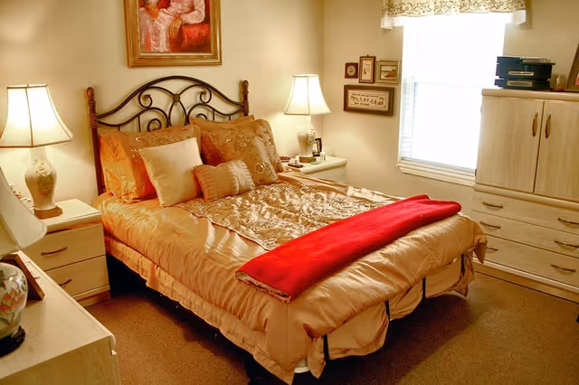 Sunlit furnished bedroom with a double bed dressed in gold bedding and a red throw, flanked by nightstands and lamps, with a dresser under the window.