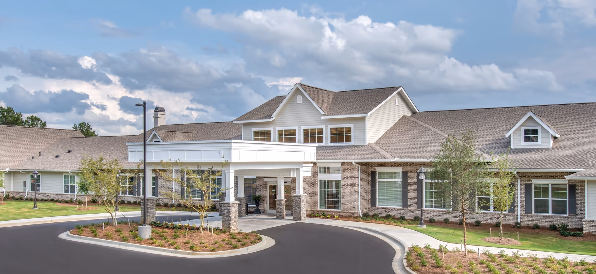 Exterior view of a single-story senior living facility with a covered entrance, brick and siding facade, multiple windows, landscaped greenery, and a curved driveway under a partly cloudy sky.
