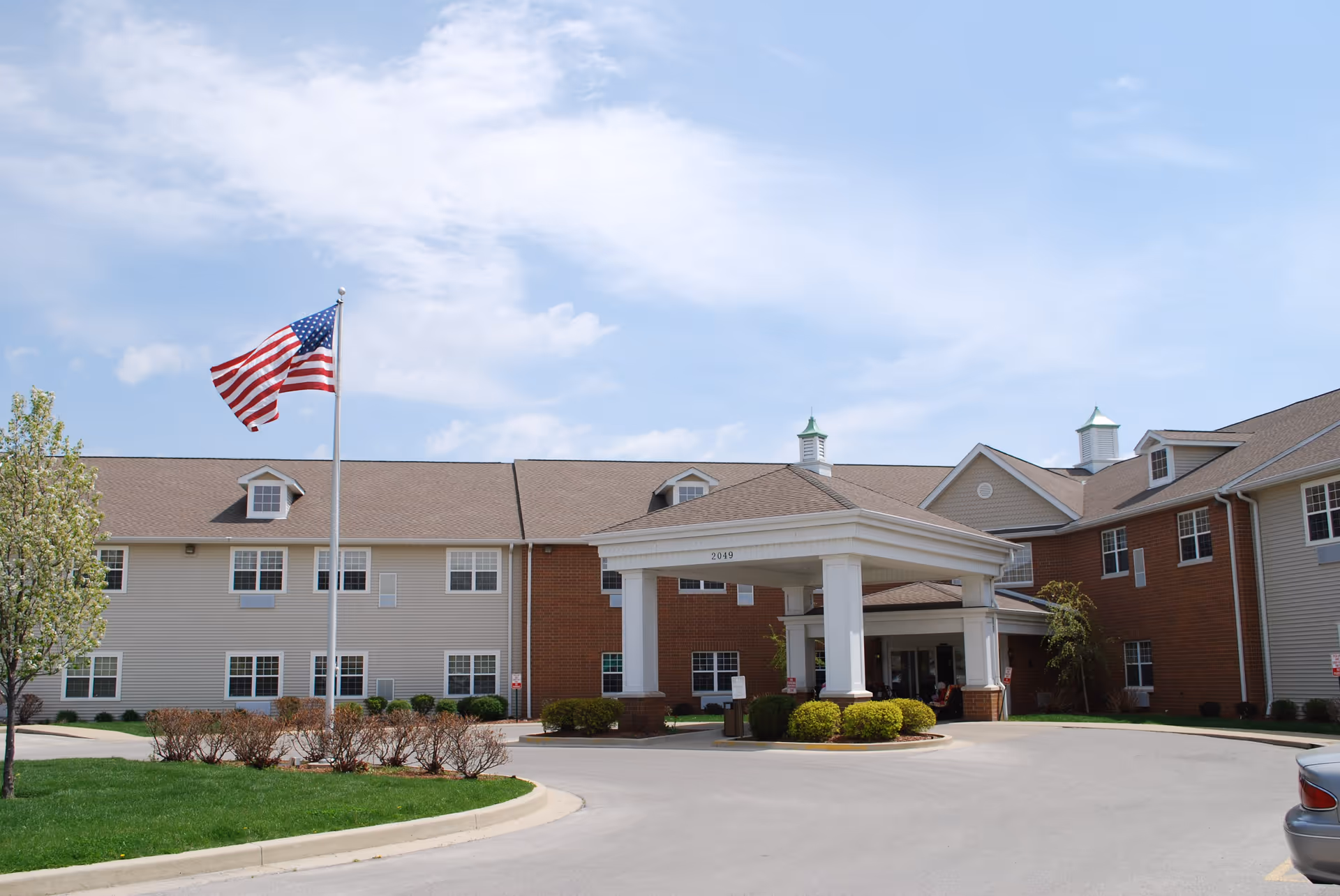 Exterior view of Heritage Woods Of Centralia, showing a two-story building with beige and red brick siding, a covered entrance with white columns, an American flag on a flagpole, and a clear blue sky with some clouds.