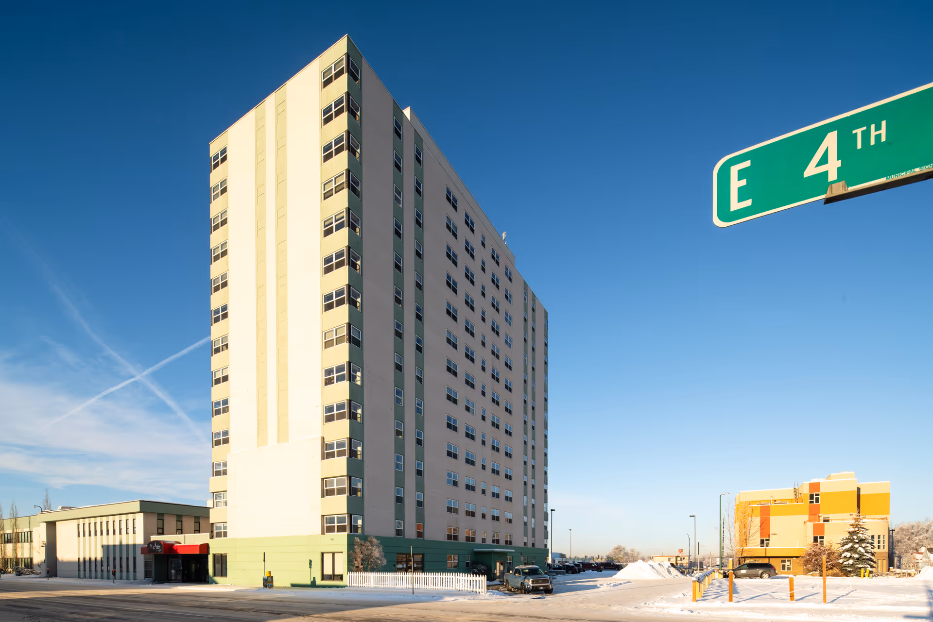 Tall multi-story building with green and beige exterior under a clear blue sky, snow on the ground, and a street sign for E 4th visible in the foreground.