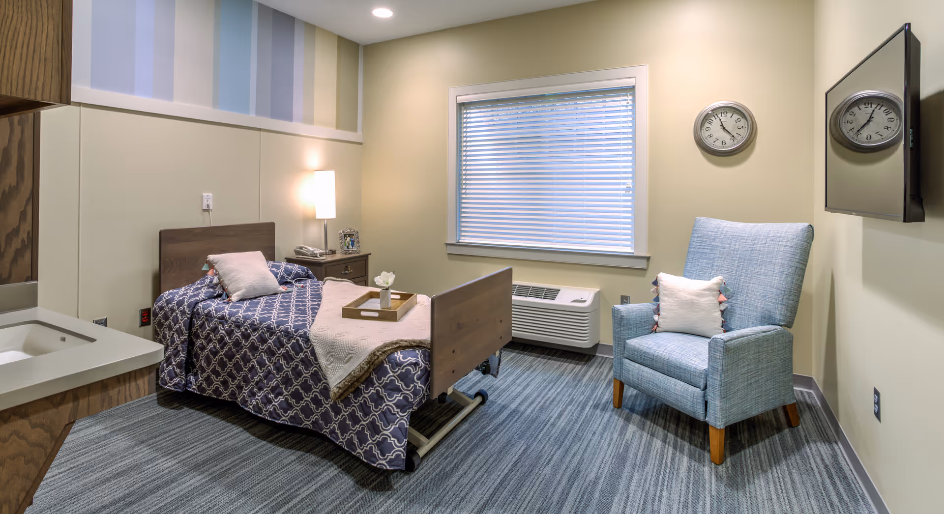 A neatly arranged nursing facility room with a single bed covered in a blue patterned bedspread and a beige throw blanket. There is a wooden nightstand with a lamp, a photo frame, and a phone next to the bed. A blue upholstered armchair with a white pillow is positioned near a window with closed blinds. A wall-mounted TV and a clock are visible on the wall. The room has a carpeted floor and a small sink area in the corner.