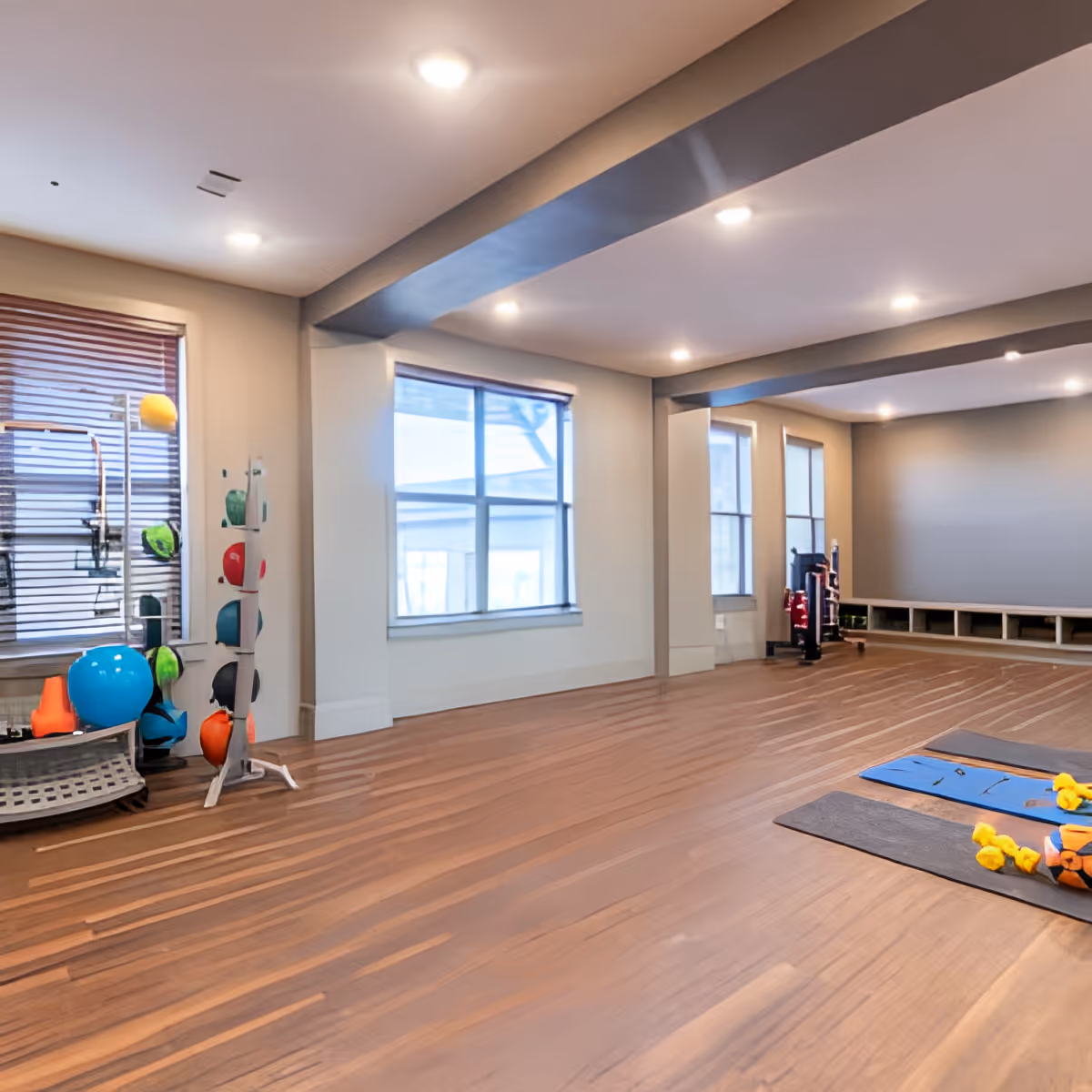 A spacious exercise room with wooden flooring and large windows letting in natural light. The room contains exercise mats with small yellow dumbbells and a basketball, a rack with various colorful medicine balls, and other fitness equipment. The ceiling has recessed lighting and structural beams.