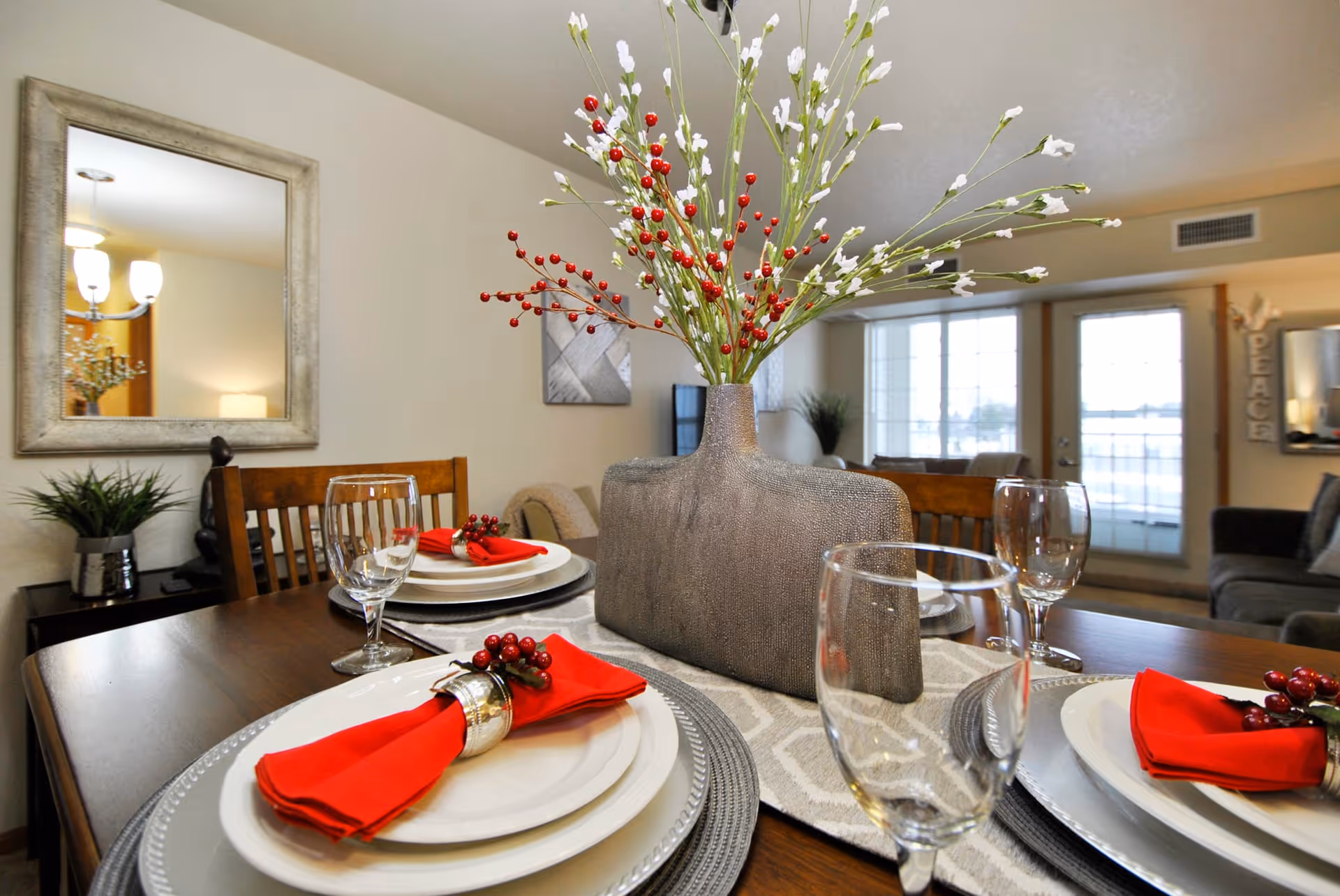 A dining table set for four with white plates, red napkins with berry decorations, and clear wine glasses. A large decorative vase with white and red artificial flowers is centered on the table. In the background, there is a living room area with a couch, a large window, and a door leading outside. A mirror and a small plant are visible on the wall to the left.
