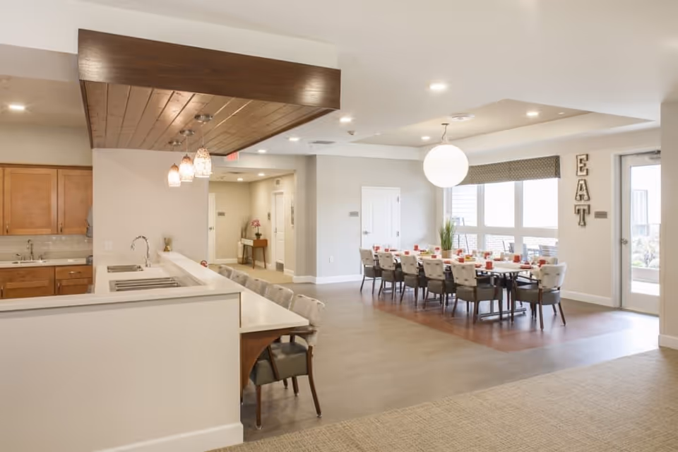 A bright and spacious dining area in a senior living facility featuring a long dining table set with red cups and plates, surrounded by cushioned chairs. To the left, there is a kitchen counter with bar stools and pendant lights hanging from a wooden ceiling accent. Large windows allow natural light to fill the room, and the word 'EAT' is displayed vertically on the wall near a glass door.