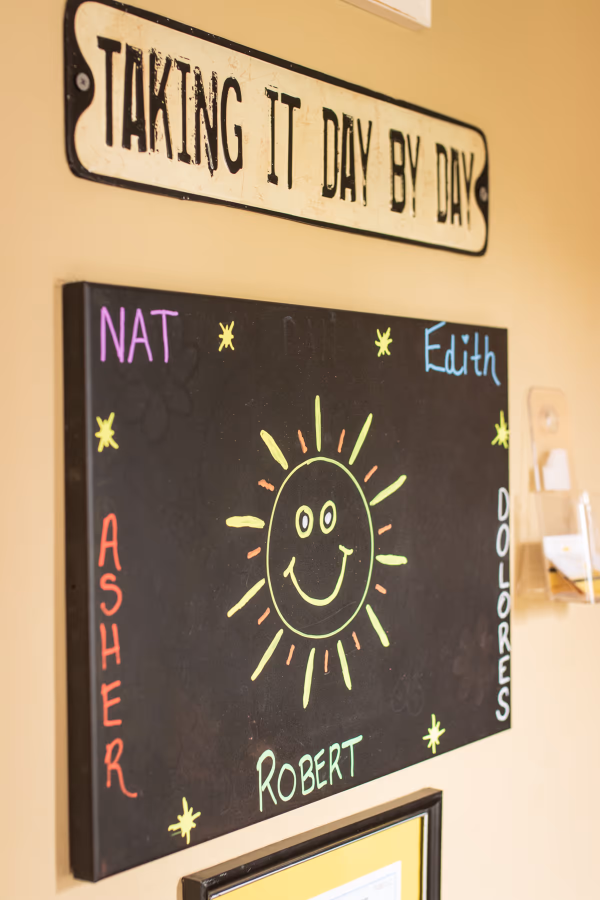 A wall with a decorative sign that reads 'TAKING IT DAY BY DAY' above a blackboard featuring a colorful drawing of a smiling sun surrounded by names written in different colors: NAT, ASHER, ROBERT, EDITH, and DOLORES.